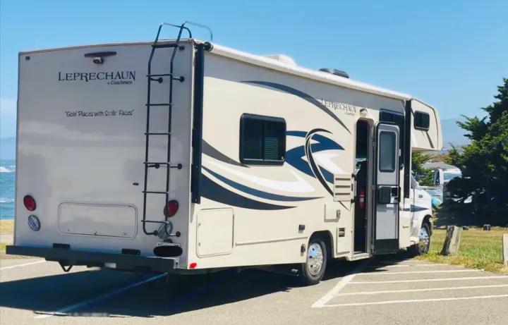 A tan Coachmen Leprechaun RV rental, parked on asphalt with its door open, facing the ocean. Perfect for a seaside road trip.. Coachmen Leprechaun 2016