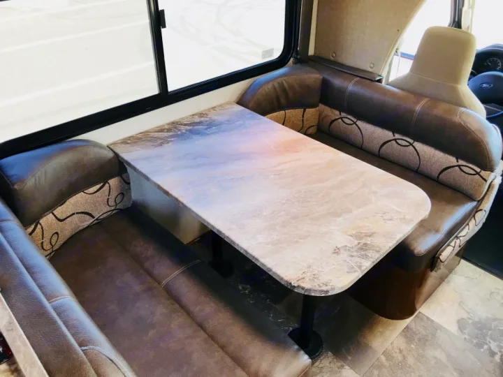 View of an RV rental dinette with brown leatherette and patterned fabric booth seating, and a light marble-patterned table.. Coachmen Leprechaun 2016