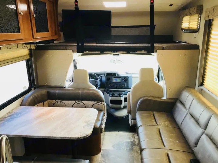 This RV rental interior shows the living space: a dinette, a sofa, and an overhead bunk above the cab area.. Coachmen Leprechaun 2016