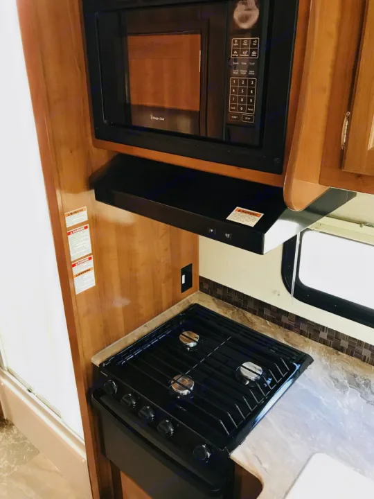 Compact RV rental kitchen: black microwave, range hood, & 3-burner gas stove. Ready for meals on the road.. Coachmen Leprechaun 2016