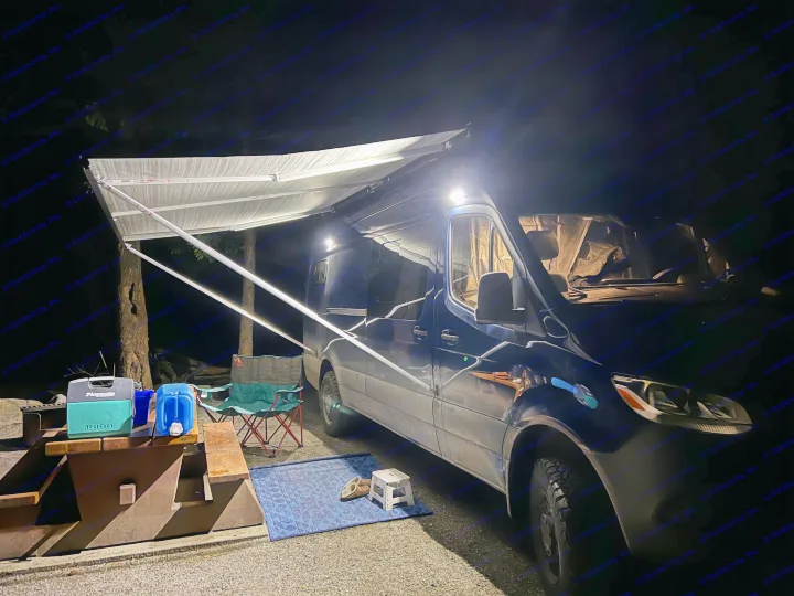 Nighttime scene: a blue camper van set up for camping. Its illuminated awning extends over a picnic table with gear. Perfect for an RV rental.. Mercedes Sprinter 2022