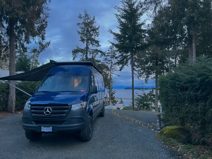 Blue RV rental parked at a scenic waterfront campsite with awning extended, offering beautiful views.. Mercedes Sprinter 2022