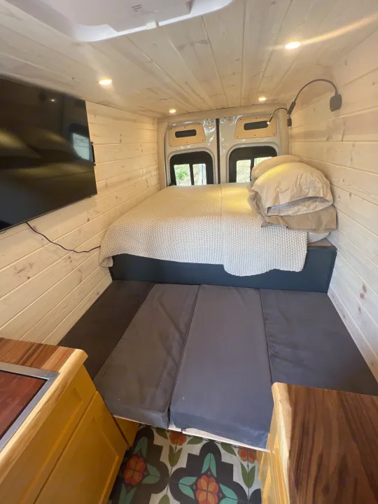 Inside an rv rental: wood-paneled walls, bright bed area, TV, and floor cushions. A comfortable mobile living space.. Mercedes-Benz Sprinter RV Motorhome Campervan 2022