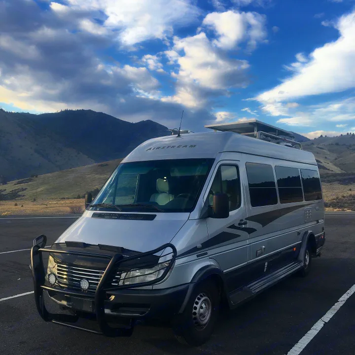 A silver Airstream Sprinter van, equipped as an RV rental with solar panels & a bull bar, parked before mountains.. Airstream Interatate 2003