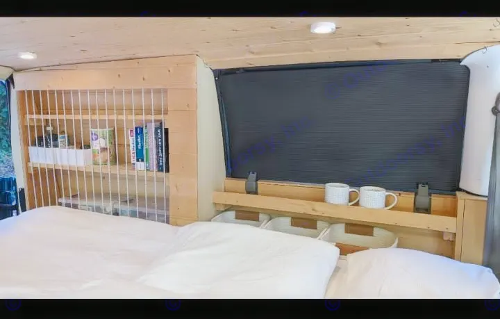 A custom RV rental interior features a cozy bed, wood shelving with books, a window with a black blind, and mugs on a ledge.. Chevrolet Other 1996