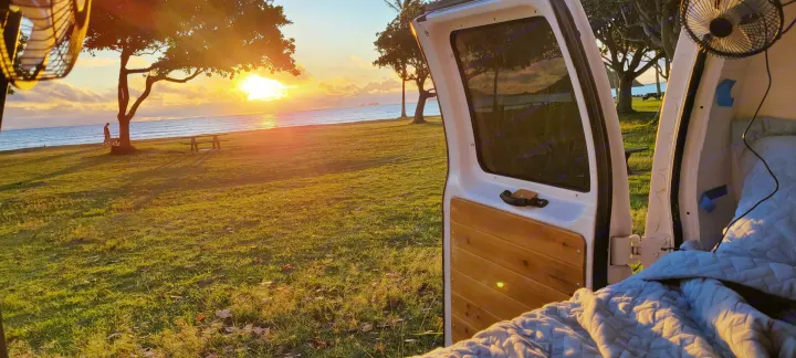 View from an RV rental's open back door: golden sunset over ocean, grassy park with a person and dog, and a cozy bed inside.. Ford E250 2009