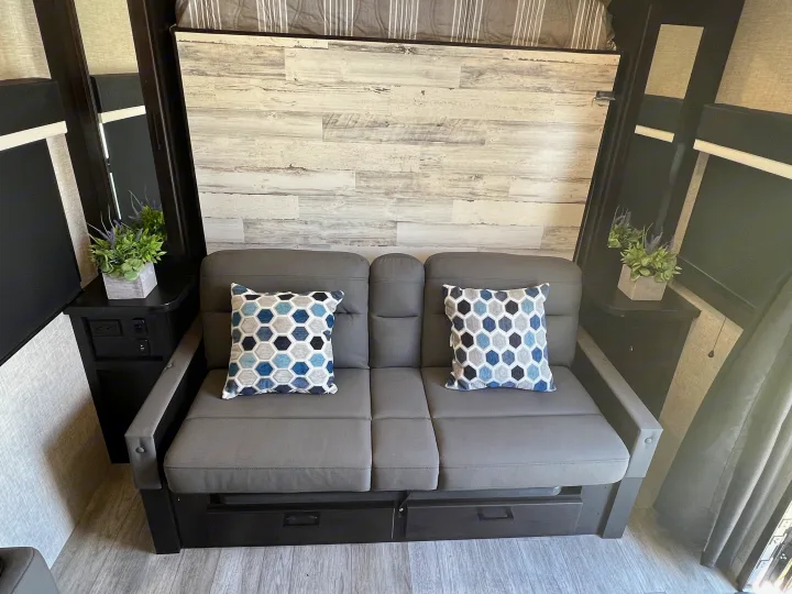 Cozy RV rental interior featuring a grey sofa with storage, blue/grey geometric pillows, and side tables with outlets.. Jayco 21MML 2023