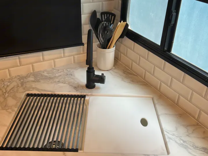 Modern RV rental kitchen: white marble-look counter, black faucet, sink with drying rack/cutting board, and tiled backsplash.. Jayco 21MML 2023