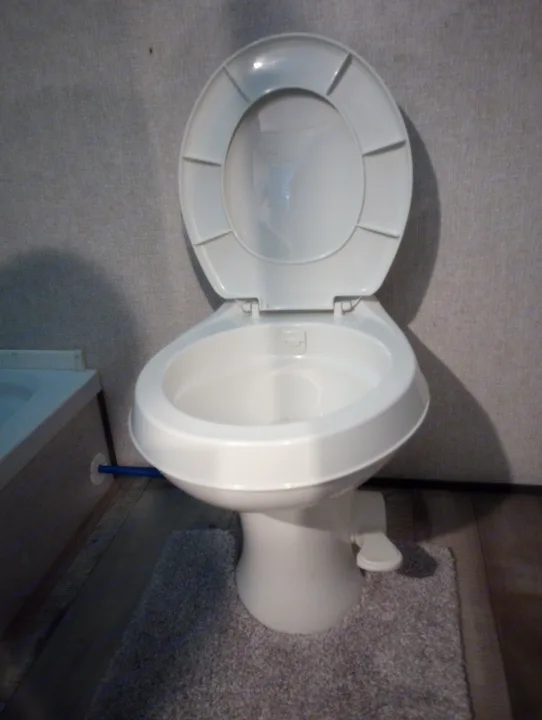 Clean white toilet with lid up in a compact RV rental bathroom, a blue water pipe visible.. Dutchmen 17BH 2021