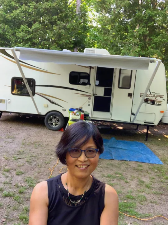 Happy woman by her Escape rv rental, a white travel trailer, with an extended awning at a wooded campground.. Escape Trailer Industries Escape 5.0ta 2013
