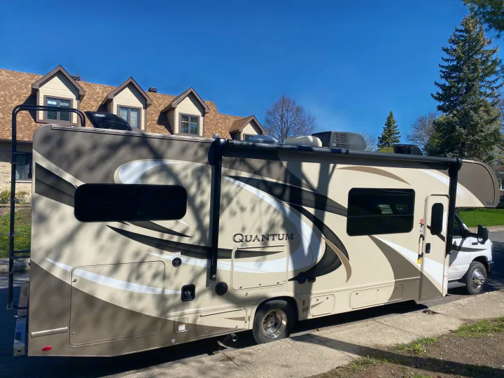 A modern tan and brown Class C RV, an ideal RV rental, parked on a sunny day in a residential area. Great for travel adventures.. Thor Quantum 2018