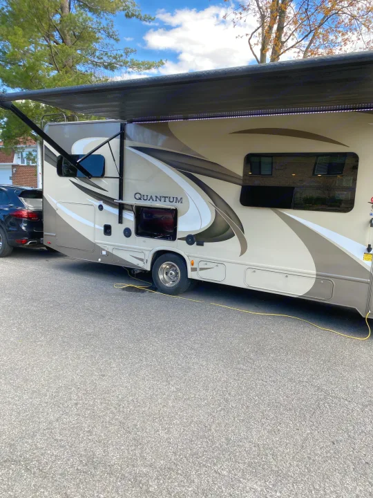 A beige and brown Quantum RV rental is parked with its awning extended, featuring an outdoor TV. A yellow cord is visible.. Thor Quantum 2018