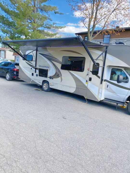 This beige and brown RV rental, a Quantum motorhome with its awning extended and lights on, is parked on a street with a black SUV hitched.. Thor Quantum 2018