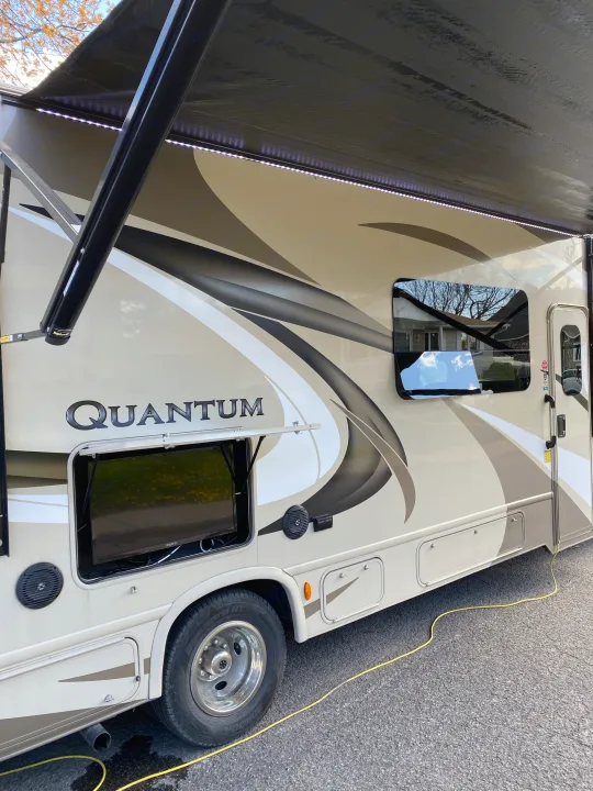 Side view of a Quantum RV rental motorhome with an extended awning, LED lights, and an open compartment revealing an outdoor TV.. Thor Quantum 2018
