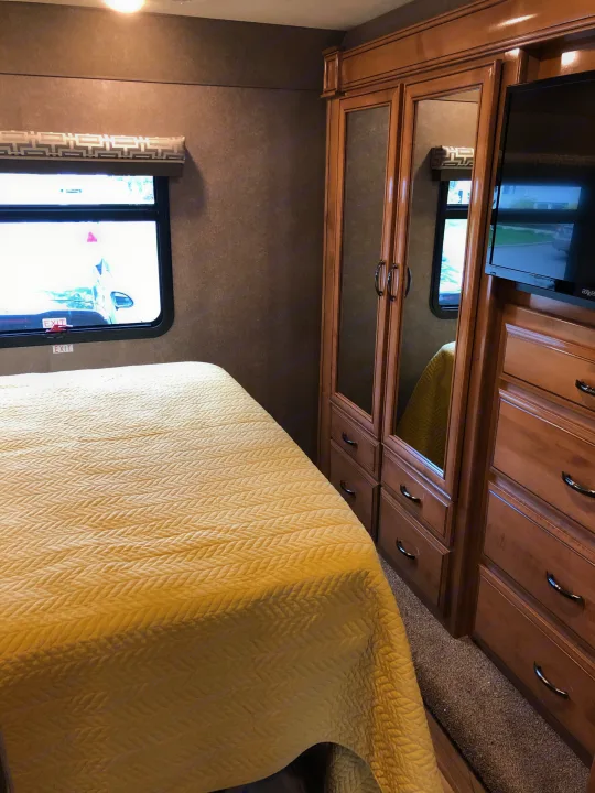 RV rental bedroom interior featuring a comfortable yellow bed, window, and wood storage cabinets with mirrors.. Thor Quantum 2018