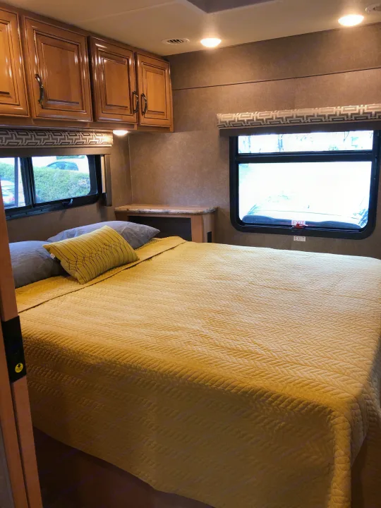 Cozy RV rental bedroom: yellow quilted bed, wooden cabinets, and windows with patterned shades.. Thor Quantum 2018