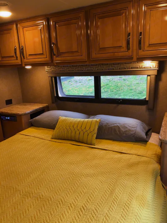 Inside an RV rental: a cozy bedroom with bright yellow quilted bedding, wood cabinets, and a scenic window view.. Thor Quantum 2018