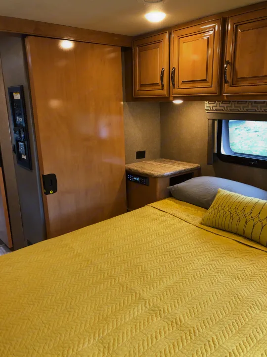 Cozy RV rental bedroom features a yellow bed, compact nightstand, and wooden overhead cabinets.. Thor Quantum 2018