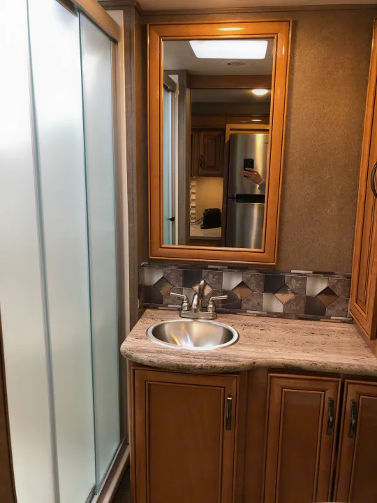 RV rental bathroom: Vanity with stainless sink, chrome faucet, and tiled backsplash. Frosted shower door visible. Mirror reflects kitchen.. Thor Quantum 2018