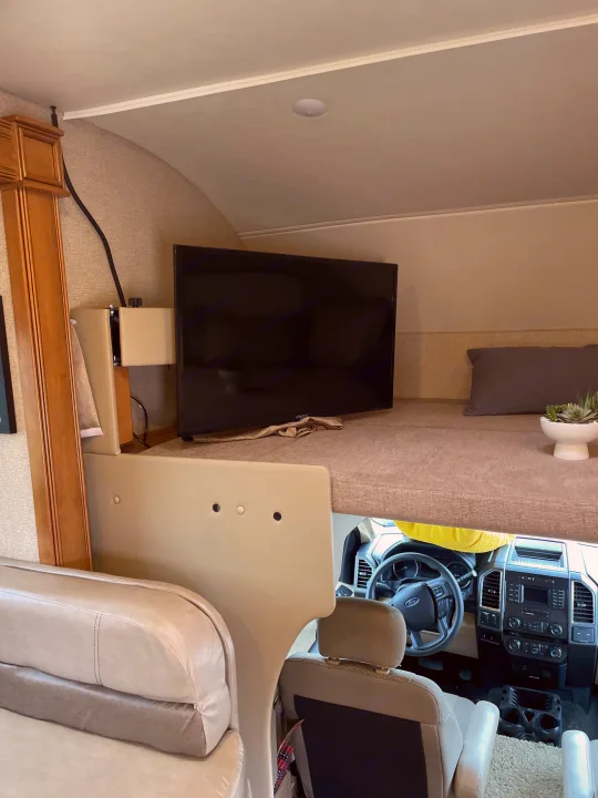 Cozy RV rental interior showcasing an over-cab bunk bed with a TV, overlooking the driver's area.. Thor Chateau 2018