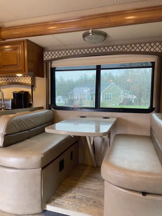 An RV rental dinette setup with plush benches, a table, and a kitchen area, looking out a rain-splattered window.. Thor Chateau 2018