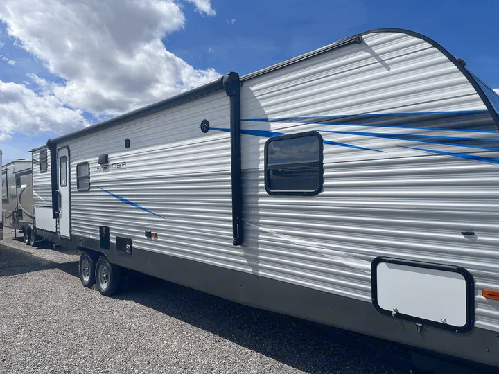 A modern Avenger travel trailer for rv rental, featuring light gray siding, blue graphics, and an awning under a cloudy sky.. Forest River Avenger 29QBS 2021