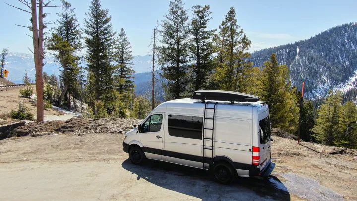 Silver camper van with roof box, perfect for an RV rental mountain road trip, parked amidst pine trees & snowy peaks.. Mercedes-Benz Sprinter 2016