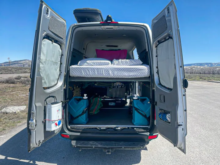 This custom rv rental features a cozy elevated bed, ample storage with water tanks, and a mounted portable shower for van life adventures.. Mercedes-Benz Sprinter 2016