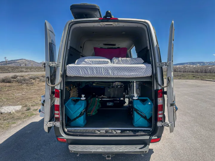 Open rear doors of an RV rental show a cozy bed, ample storage, and water containers, ready for adventure in the mountains.. Mercedes-Benz Sprinter 2016