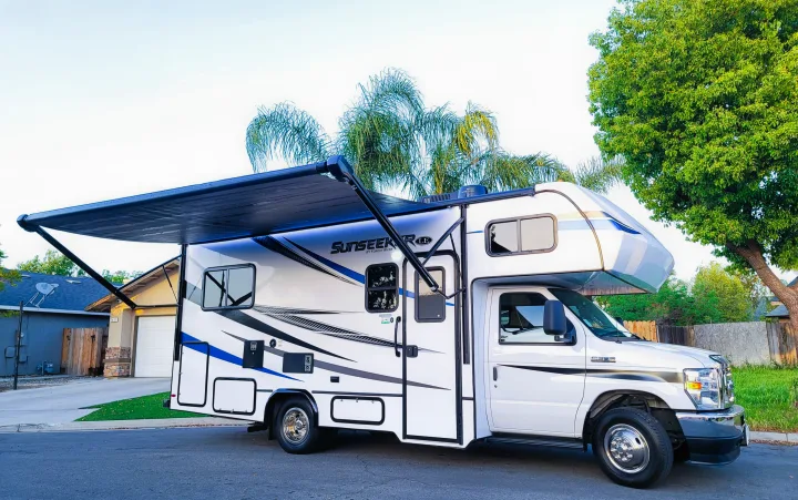 A spacious white RV rental motorhome with an extended black awning is parked on a suburban street, ready for travels.. Forest River Sunseeker LE 2022