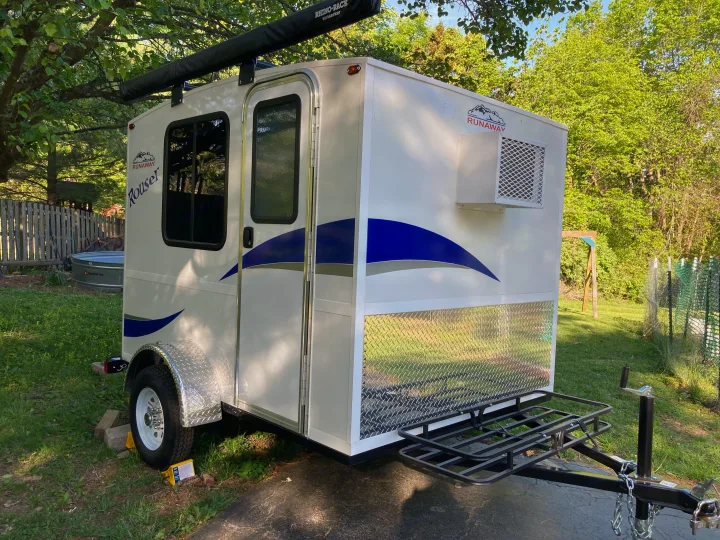 A white compact Runaway Rouser RV rental teardrop camper, featuring an awning, AC, and front cargo rack, parked on asphalt.. Runaway Rouser 2023