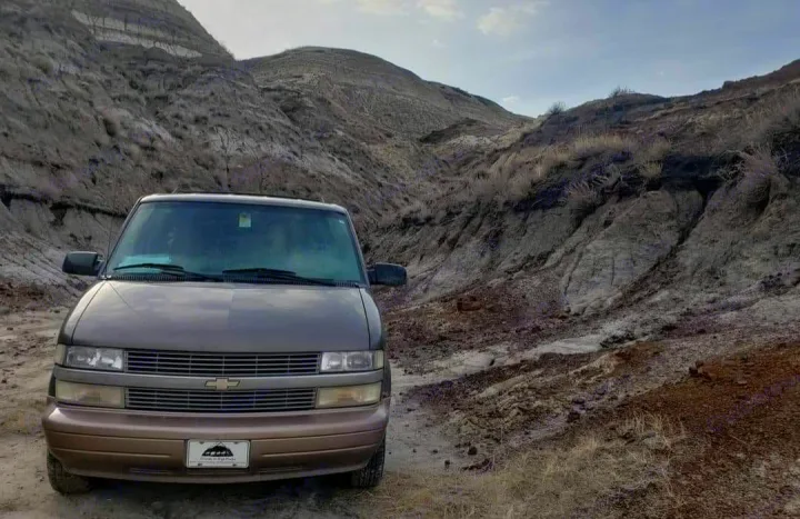 Brown Chevrolet van with RV rental plate on dirt path in rugged, dry badlands. Ideal for adventuring!. Chevrolet Open Road 2000