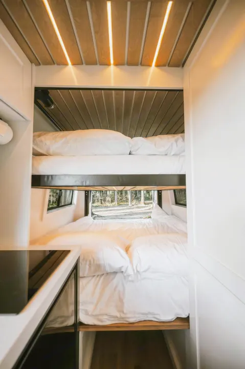 Inside a modern RV rental: stacked bunk beds with white linens, ambient wood ceiling lighting, and a window view of a serene forest.. Mercedes-Benz Sprinter 2021