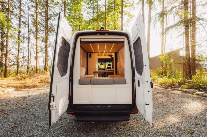 White camper van RV rental with back doors open, revealing a lit, wooden interior & grey cushions. Parked on gravel, surrounded by trees.. Mercedes-Benz Sprinter 2021