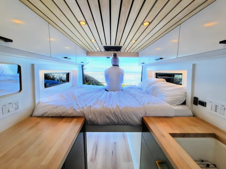 Inside a bright **rv rental**, a person enjoys an ocean view from a large bed in a modern camper van.. Ram Promaster 2500 2021