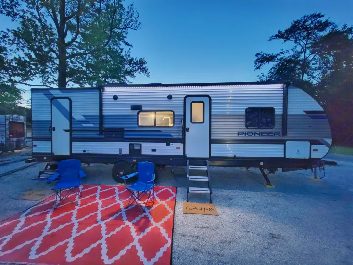 Cozy RV rental setup: a travel trailer at dusk with bright awning lights, a red outdoor rug, and blue camping chairs.. Heartland RVs Pioneer 2021