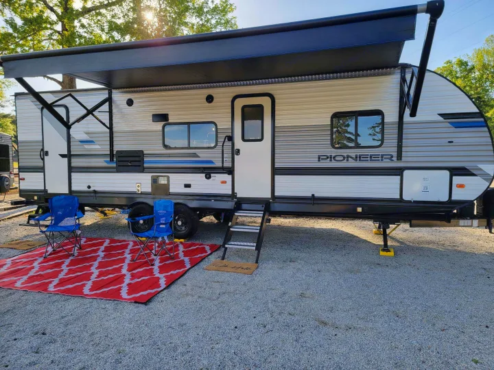An RV rental set up with an extended awning, outdoor chairs, and a red and white mat on a gravel campsite.. Heartland RVs Pioneer 2021