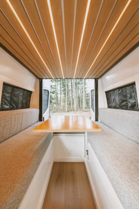 RV rental interior features: wood ceiling with linear LEDs, comfy grey benches, and a dining table. Rear doors reveal a serene forest view.. Mercedes-Benz Sprinter 2021