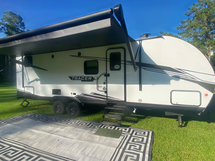White travel trailer RV rental with black awning deployed over a patterned outdoor mat on green grass. Perfect for camping.. Other Tracer 2022