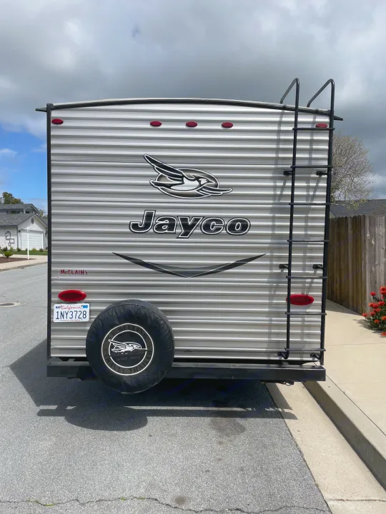 Rear view of a parked Jayco travel trailer, likely an RV rental, showing its spare tire, ladder, and California license plate.. Jayco Jaycoflight 2017
