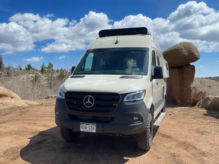A tan Mercedes-Benz Sprinter rv rental van is parked on a dirt path, surrounded by large boulders under a cloudy blue sky.. Winnebago Revel 2022