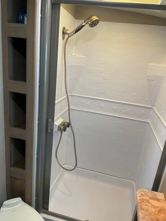 RV rental's compact bathroom features a clean white shower stall with a handheld showerhead, next to storage shelves.. Grand Design Imagine 2022