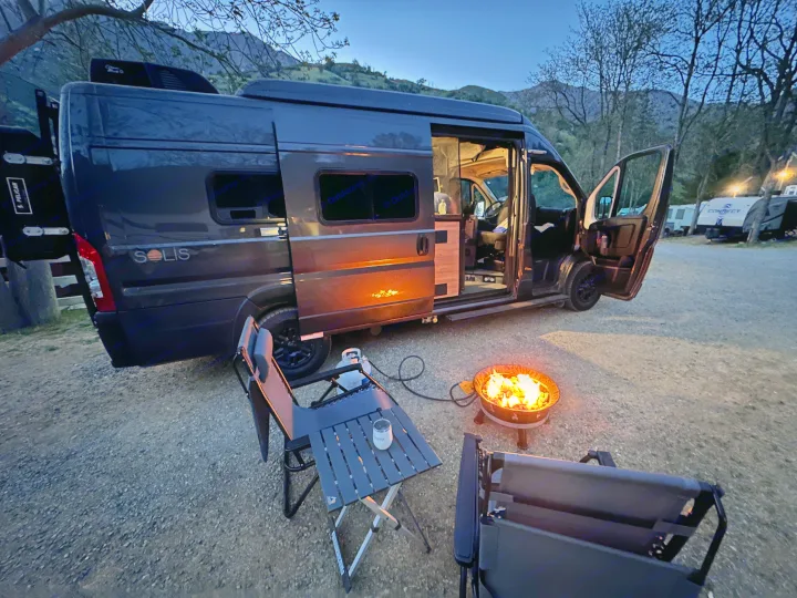 Solis RV rental parked at dusk in a mountain campsite, with a cozy propane campfire, table, and chairs set up for relaxation.. Winnebago Solis 2022
