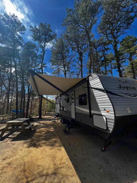 A spacious RV rental travel trailer with an extended awning is parked at a sunny wooded campsite, with a picnic table nearby.. Jayco Jay Flight SLX 2020