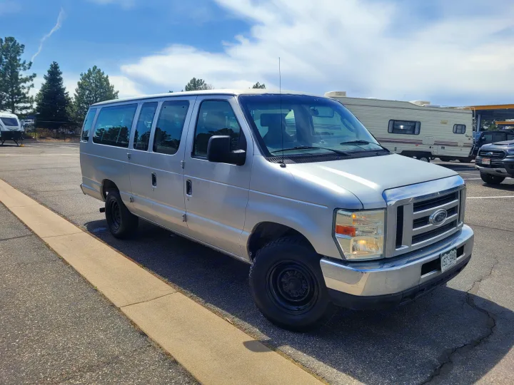 A silver Ford van is parked on asphalt at an RV rental lot, with a white travel trailer and dark truck in the background.. Ford Ford E350 2009