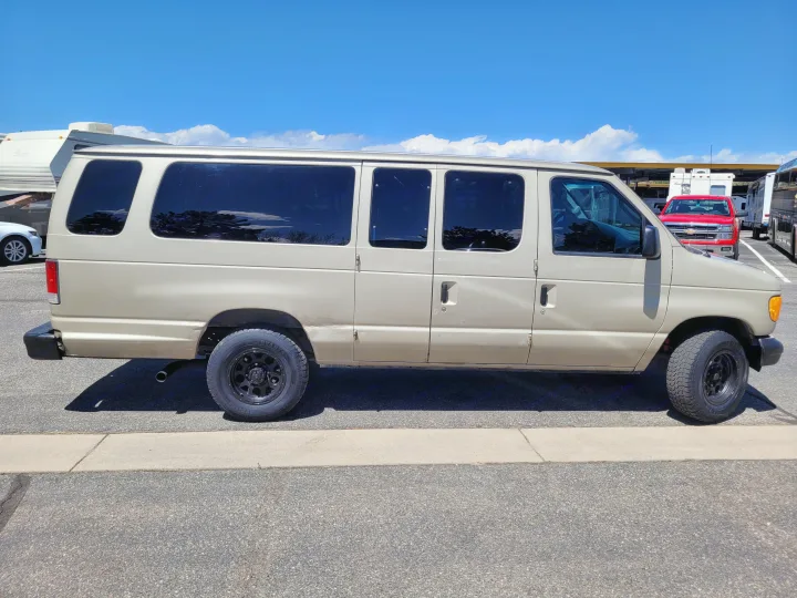Tan Ford van with all-terrain tires, possibly an RV rental service vehicle, parked next to RVs under a blue sky.. Ford E-350 2007