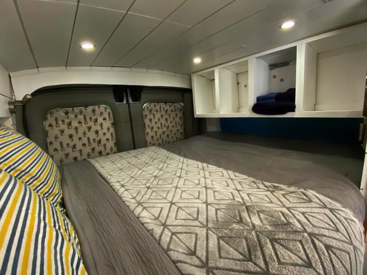 RV rental's cozy interior: bed with patterned blanket, overhead storage, and cactus window curtains. Perfect for travel.. Ford Transit Custom 2015