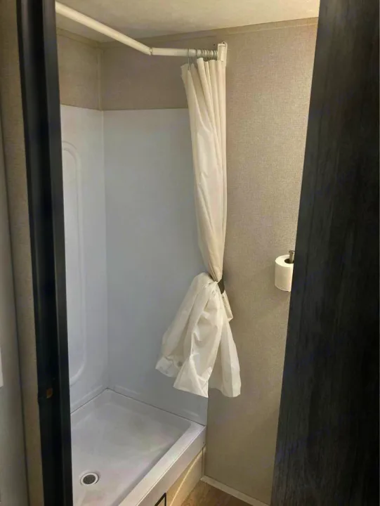 RV rental compact white shower: white curtain on L-shaped rod, gathered, with toilet paper dispenser.. Keystone RV Hideout 2022