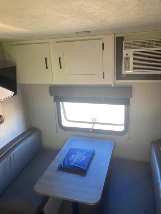 This RV rental's dinette area features a light-colored table, dark grey bench seating, white overhead cabinets, and an AC unit.. Keystone RV Hideout 2022