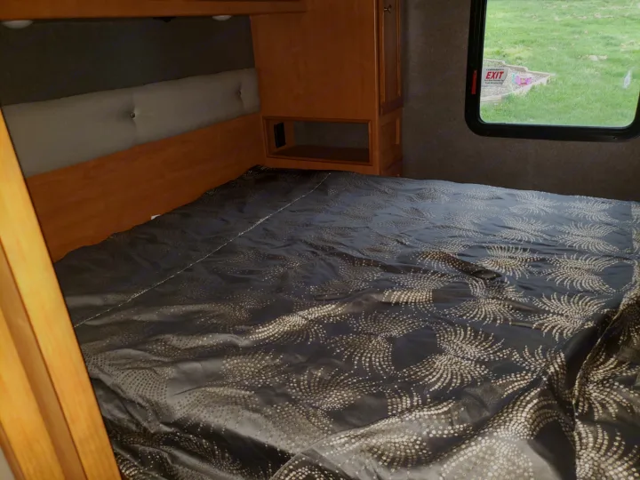 RV rental bedroom: Cozy bed with dark patterned comforter, wood cabinets, window view. Great for trips!. Winnebago Vista 2017